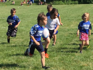 Six children running on a green grass field. Four children are wearing blue rugby uniform shorts. Two Children wearing white t-shirts. The child in the foreground is carrying the ball in one hand. His head is held high as he scans the defense. The other children are chasing the ball carrier.
