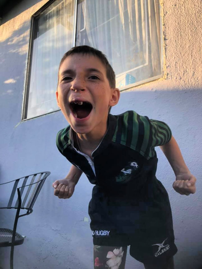 A young child wearing a black rugby jersey with green hoops on the sleeve and black rugby shorts over green flower print tights. The child has short black hair. The child is lunging forward, fists cocked back at the waist, face showing an intense roar.