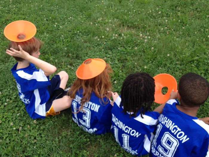 Four children sitting in a grass field. Seen from behind. Each child wears a blue jersey with white lettering that reads, Washington and white numbers. Each of the first two children from left to right has an orange soccer cone on their head. The third child has a cone in their hands. The fourth child is looking at the first three.