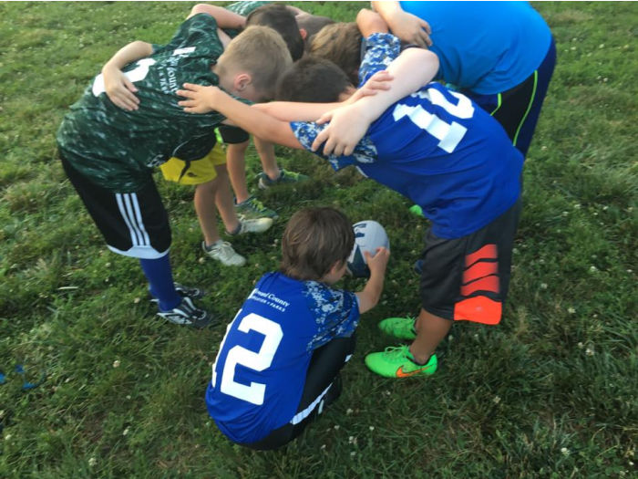 Six children engage in a scrum. The scrum is unbalanced with three players in green jerseys on the left and two players in blue jerseys on the right. These five are bent over facing each other and bound together with their arms around each other. The sixth child, in a blue jersey, places the ball in the tunnel formed by the other five players