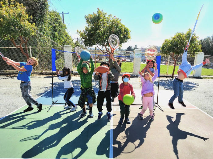 kids on tennis court with football, basketball, and tennis rackets