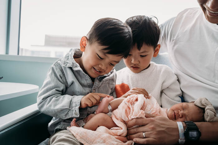 two big kids and their newborn sibling touch the baby's toes at the hospital