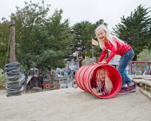 two girls playing at adventure playground berkeley. one is rolling the other around in a tube
