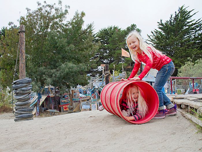 two girls playing at adventure playground berkeley. one is rolling the other around in a tube
