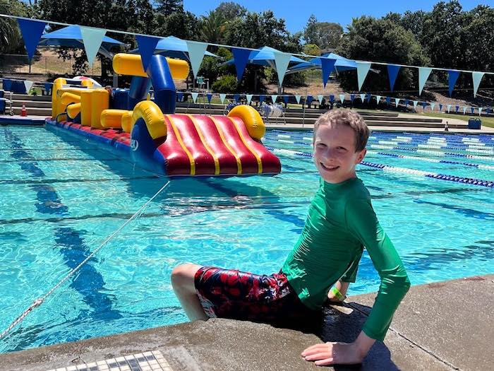 child smiling at swimming pool in front of inflatable obstacle course that floats at the pool