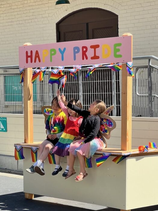 3 children at the bay area discovery museum wearing rainbows and carrying pride flags on a booth that reads happy pride