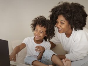 Black adult and child wearing white shirts and jeans use a laptop together for a video call or funny video