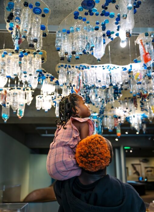 child on adults shoulders in the science gallery under an artwork of plastic bottles and trash