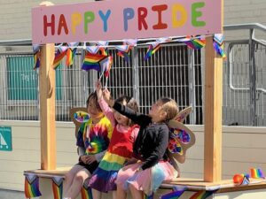 3 girls on a HAPPY PRIDE booth wearing rainbow clothing and waving flags