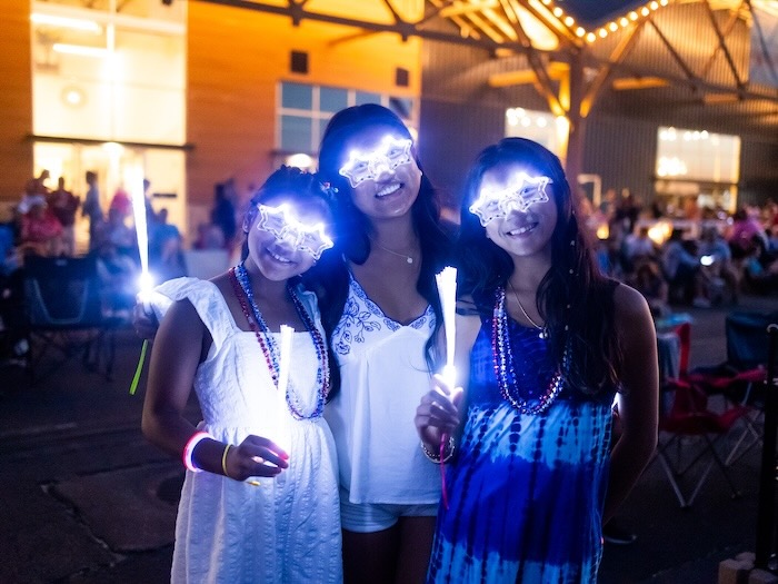 three teen girls holding light up toys and wearing light up star glasses celebrating fourth of july