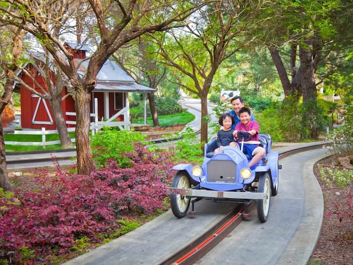 family of two kids and one adult riding in an old timey car on a paved track at gilroy gardens