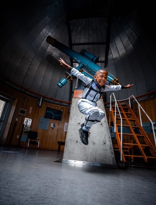 child jumping while wearing a space suit in front of the telescope