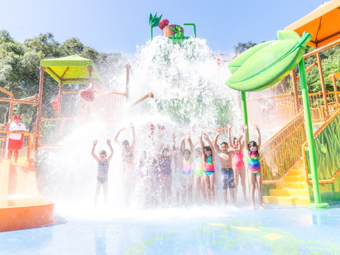 children under a water feature at a sprayground wearing swim suits near a water slide