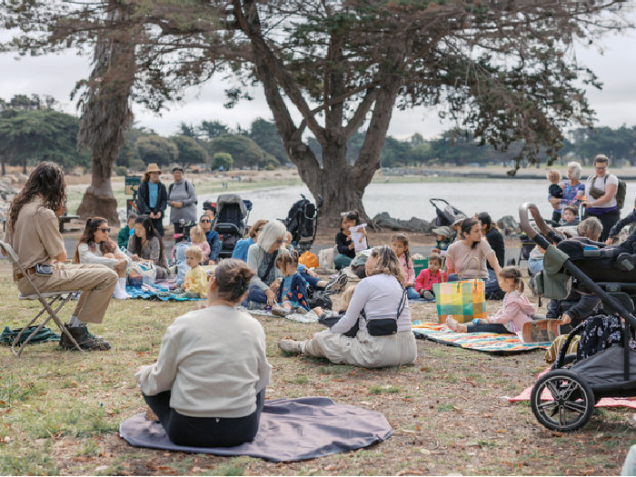 lots of kids and adults sitting on blankets listening to a story at the water's edge